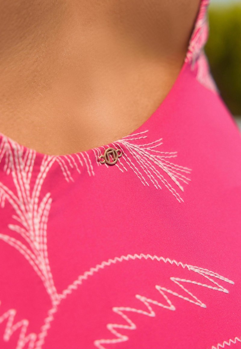 Lys pink stof med hvide broderede palme mønstre og en metal logo detalje nær halsudskæringen. Glat tekstur, tætsiddende design.