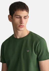 T-shirt in cotone verde con scollatura rotonda, maniche corte e un piccolo logo ricamato sul petto. Texture liscia, design aderente.