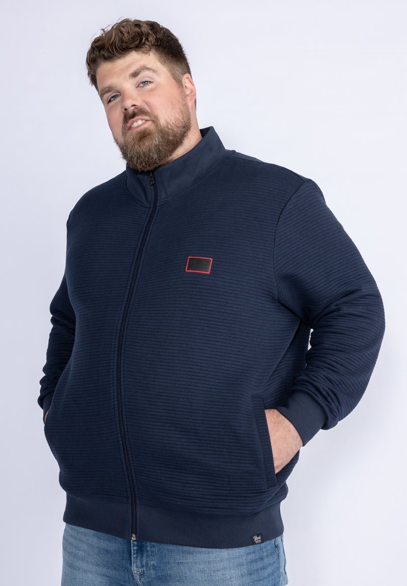 Navy blue zip-up jacket with ribbed texture, high collar, side pockets, and a red accent label. Casual design suitable for layering.
