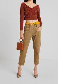 Rostfarbenes langärmliges Oberteil mit gerafftem Vorderteil, beigefarbene Hose mit Taschen, gelber Gürtel, braune Tasche mit Krokodil-Textur und Schlangenprint-Absätze.
