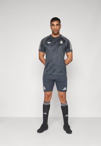 Maglia sportiva grigio scuro con motivo geometrico, tre strisce bianche sulle spalle, logo sul petto, abbinata a pantaloni shorts coordinati e calze al ginocchio.