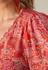 Rote Bluse mit floralem Paisleymuster, rundem Ausschnitt, Knöpfen und puffärmel. Leichter, strukturierter Stoff mit bunten Akzenten.