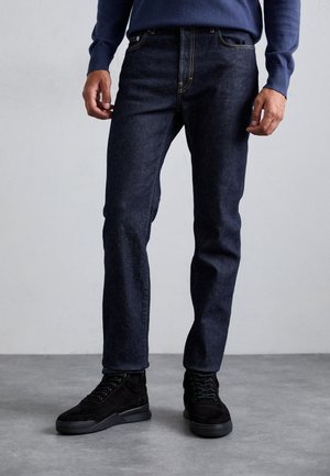 Hombre con jeans azul oscuro y zapatillas negras de pie sobre un suelo gris frente a una pared lisa de color gris claro.