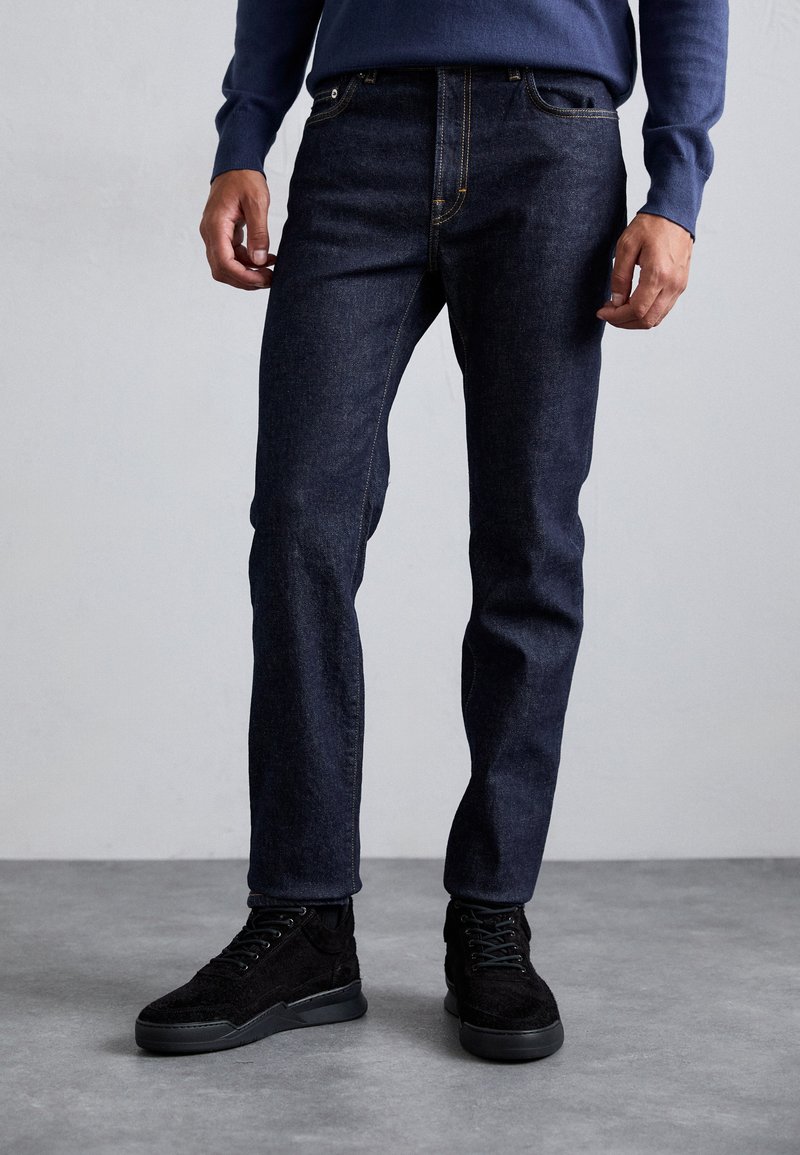 Homme portant un jean bleu foncé et des baskets noires, debout sur un sol gris contre un mur gris clair uni.