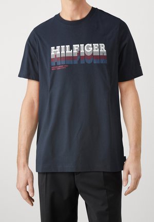 Man draagt een marineblauw T-shirt met korte mouwen met vetgedrukte witte, rode en blauwe tekst "HILFIGER Established 1985 New York City" op de borst.