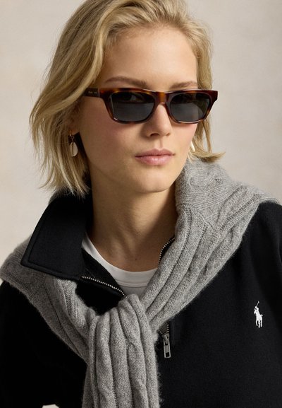 Giovane con capelli biondi corti, indossa occhiali da sole tartarugati, un top nero con zip e un maglione grigio a maglia drappeggiato sulle spalle.
