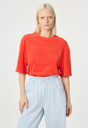 Blonde woman wearing loose red short-sleeve top and light blue high-waist pants, standing against a plain light background.