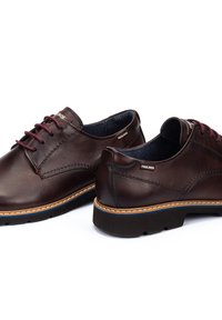 Zapatos de cuero marrón con punta redonda, costuras en contraste y cordones burdeos. Presentan una suela de goma y un detalle de logo grabado en el lateral.
