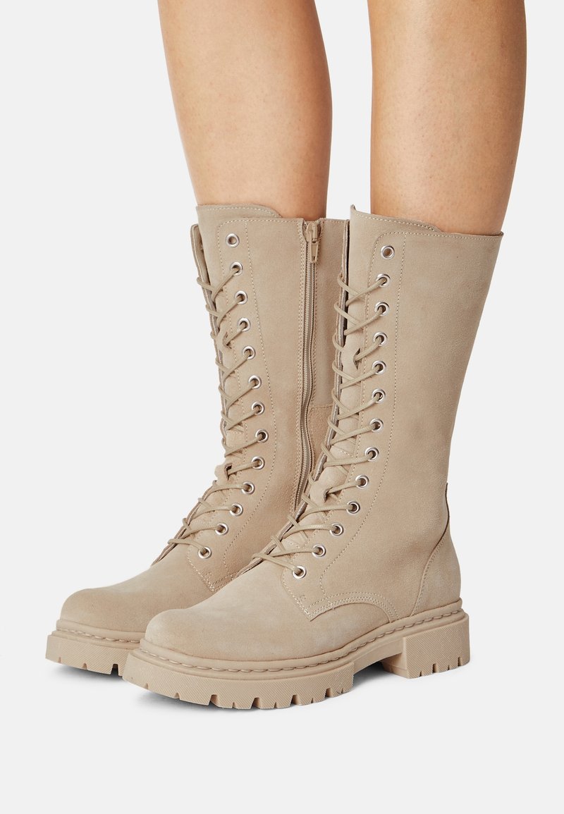 Beige knee-high boots made of suede with a lace-up front, zipper closure, and rugged rubber sole featuring prominent tread and stitching details.