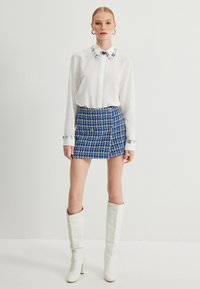 Witte button-up shirt met bloemenkraag, gecombineerd met een blauwe tweed minirok met gouden knopen, en kniehoog witte haklaarzen.