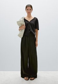 Zwart, kort mouw shirt met knopen, gecombineerd met brede, geplooide zwarte broek. Een lichtgekleurde, gestructureerde tas wordt onder de arm gedragen.