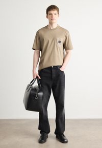 Tricou din bumbac bej cu buzunar pe piept și logo, purtat cu pantaloni din denim negri și pantofi din piele negri. Poartă o geantă din piele neagră.