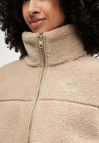 Μπεζ φλις μπουφάν με ψηλό γιακά και μπροστινό φερμουάρ, διαθέτοντας υφή μαλακού υφάσματος και μια μικρή λεπτομέρεια λογότυπου κοντά στο φερμουάρ.