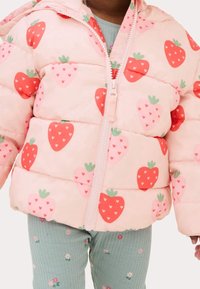 Roze pufferjack met een aardbeienpatroon, ritsluiting en een capuchon. Draagt over groene geribbelde leggings met bloemdecoraties.