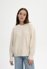 Beige Strickpullover mit strukturiertem Rippmuster, Rundhals-Ausschnitt und überschnittenen Schultern, kombiniert mit hellblauen, geraden Jeans.