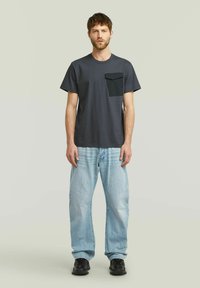 T-shirt corto grigio scuro con tasca nera a contrasto, abbinato a jeans baggy azzurri chiari. Design semplice, materiale in cotone.