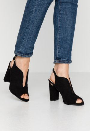 Czarne zamszowe sandały na obcasie z odkrytymi palcami i wycięciami po bokach. Gruby obcas typu blok oraz regulowany pasek wokół kostki ozdobione złotymi akcentami.