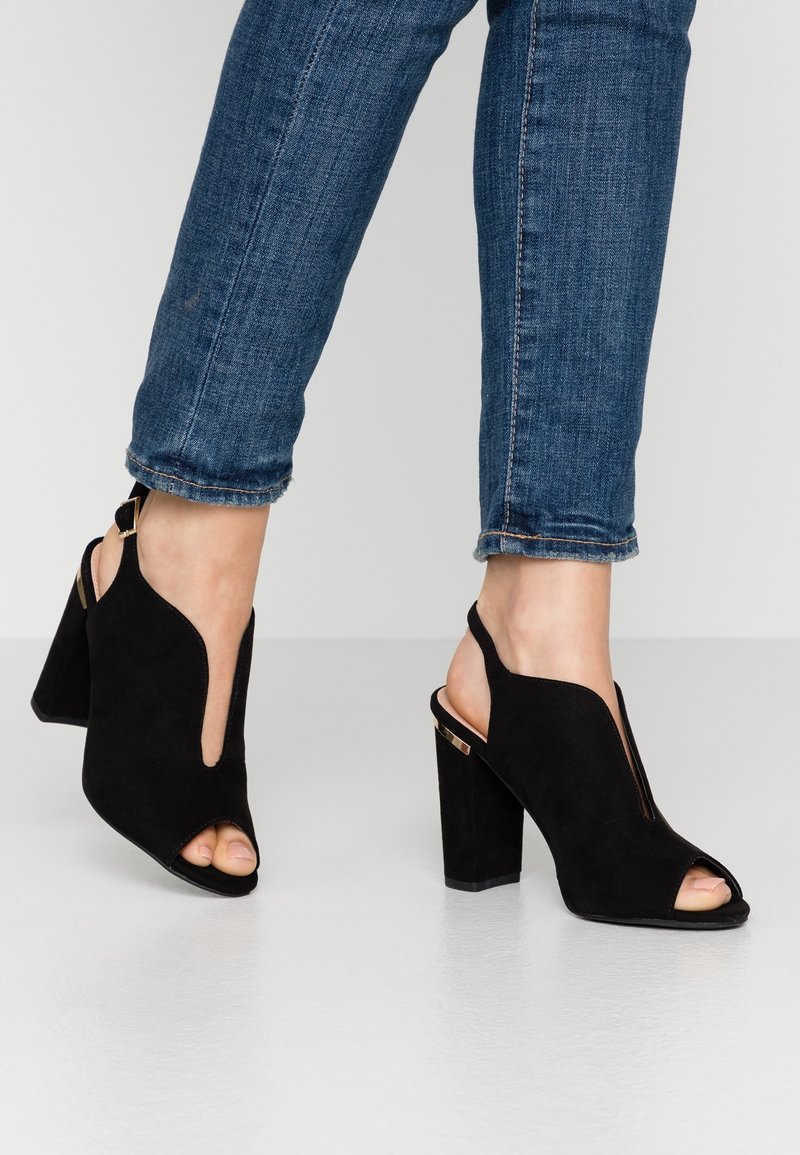 Sandales à talons en daim noir avec bout ouvert et fentes sur les côtés. Talon bloc épais et bride de cheville ajustable ornés d'accents dorés.