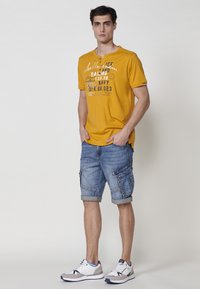 Geltona trumpomis rankovėmis marškinėliai su atspausdintais akcentais; džinsiniai cargo šortai su apvadu; pilki ir balti sportiniai batai su mėlynais ir raudonais akcentais.