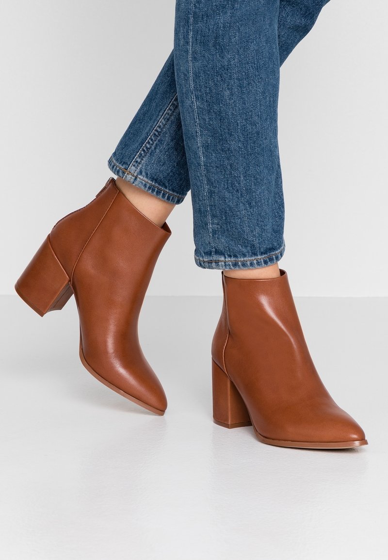 Bottines en cuir marron à bout pointu et talon bloc. Texture lisse avec une fermeture éclair latérale, portées avec un jean bleu retroussé.