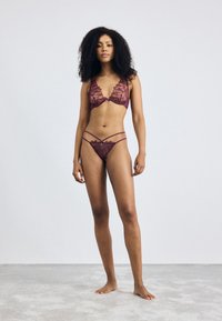 Burgundijas mežģīņu apakšveļa ar ziedu rakstiem, caurspīdīgām krūštura cupām un striķu detaļām uz bikini apakšdaļas. Modelis stāv uz mīksta pelēka pamata.
