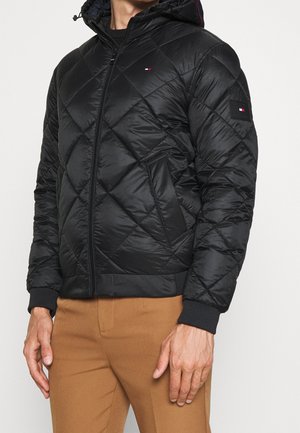 Muž v černé prošívané bundě s kapucí, na přední straně se zapínáním na zip a s malým bílým a červeným logem na hrudi a nášivkami na rukávech.