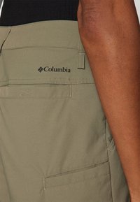 Alyvuogių žalios spalvos krovinių kelnės, pagamintos iš lengvos medžiagos, su užpakaline kišene ir juodu Columbia logotipu viršutiniame dešiniajame šone.