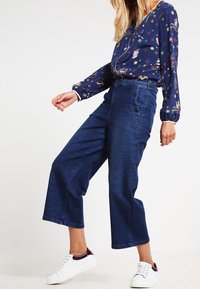 Pantaloni a gamba larga in denim scuro con vita alta, caratterizzati da dettagli a bottoni sul lato, abbinati a una blusa a maniche lunghe con motivo e sneakers bianche.