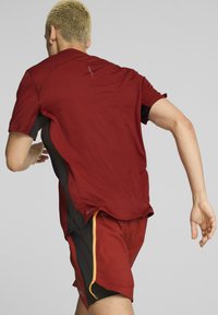Camisa atlética color burdeos con tejido texturizado, mangas cortas y paneles laterales negros; combinada con shorts burdeos a juego que presentan detalles en amarillo en la cremallera.