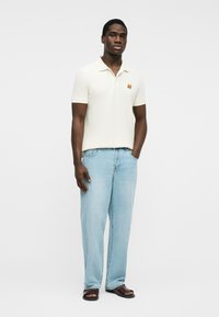 Hombre vestido con polo blanco de manga corta con pequeño logo de zorro, jeans azul claro y sandalias marrones, de pie frente a un fondo blanco liso.