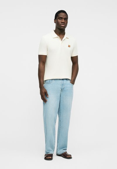 Homme portant un polo blanc à manches courtes avec un petit logo renard, un jean bleu clair et des sandales marron, debout devant un fond blanc uni.