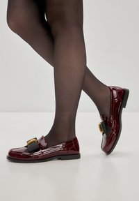 Mocassini in pelle verniciata bordeaux con una fascia nera decorativa e una fibbia dorata, caratterizzati da una punta arrotondata e un piccolo tacco nero.