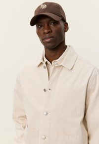 Μπεζ βαμβακερό μπουφάν με μπροστινό κούμπωμα και τσέπη στο στήθος, με ασημένια κουμπιά· συνδυασμένο με καφέ καπέλο που φέρει λογότυπο.