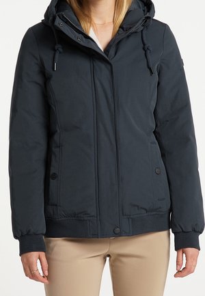 Femme portant une veste d'hiver bleu marine à capuche avec poches avant, fermeture par boutons et fermeture éclair, associée à un pantalon beige.