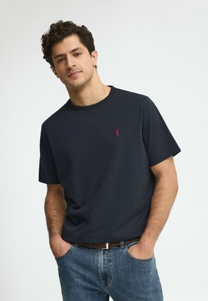 Uomo che indossa una maglietta a maniche corte blu navy scuro con logo rosso, jeans blu, in piedi con le mani nelle tasche davanti a uno sfondo chiaro e uniforme.