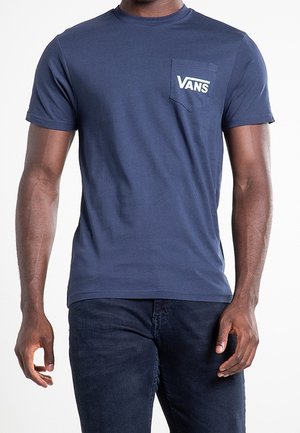 Homme portant un t-shirt Vans bleu marine avec logo sur la poche avant et un jean foncé, debout les bras détendus le long du corps.