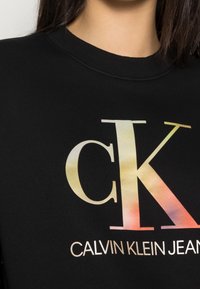 Felpa nera con un grande logo "CK" sfumato in tonalità di rosa, giallo e verde, con il testo "CALVIN KLEIN JEANS" in basso.