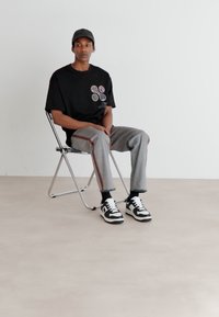 T-shirt nera con design circolari, pantaloni grigi con strisce laterali, sneakers bianche e nere, seduto su una sedia pieghevole contro un muro semplice.
