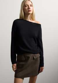 Maglione lavorato a maglia nero con spalle scoperte abbinato a una gonna di lana marrone con due tasche anteriori. Texture morbide e design aderente.