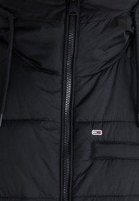 Czarna ocieplana kurtka z przodu z centralnym zamkiem błyskawicznym, mała kieszeń po prawej stronie z prostokątną naszywką z logo w kolorach czerwonym, białym i niebieskim.