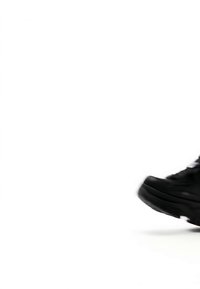 Crna tenisica usred koraka na jednobojnoj bijeloj pozadini, prikazuje đon i djelomični bočni prikaz s vidljivim vezicama.