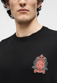 Άνδρας με μαύρο πουκάμισο λαιμόκοψης και περίτεχνο κόκκινο και λευκό κεντητό έμβλημα με την επιγραφή "Cavalli Uomo" στο στήθος, εμφανίζοντας το κάτω μέρος του προσώπου και τον λαιμό.