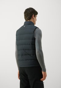 Tamnozelena puffer prsluk s horizontalnim prešanim dijelovima, stojeći ovratnik i mala oznaka na stražnjoj strani, nosi se preko sive majice dugih rukava.