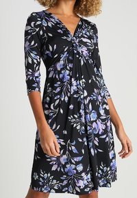 Robe noire avec un motif floral dans des tons violets et bleus. Dotée d'un décolleté en V, de manches trois-quarts et d'un design de corsage froncé.