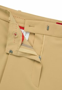 Beige broeken met een ritsgulp en knoopsluiting, met rode accenten aan de binnenkant van de tailleband en contrasterende stiksels.