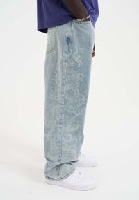 Lichtblauwe denim jeans met een losse pasvorm, voorzien van een subtiel drakenpatroon. Gecombineerd met witte sneakers, die de opgerolde zoom benadrukken.