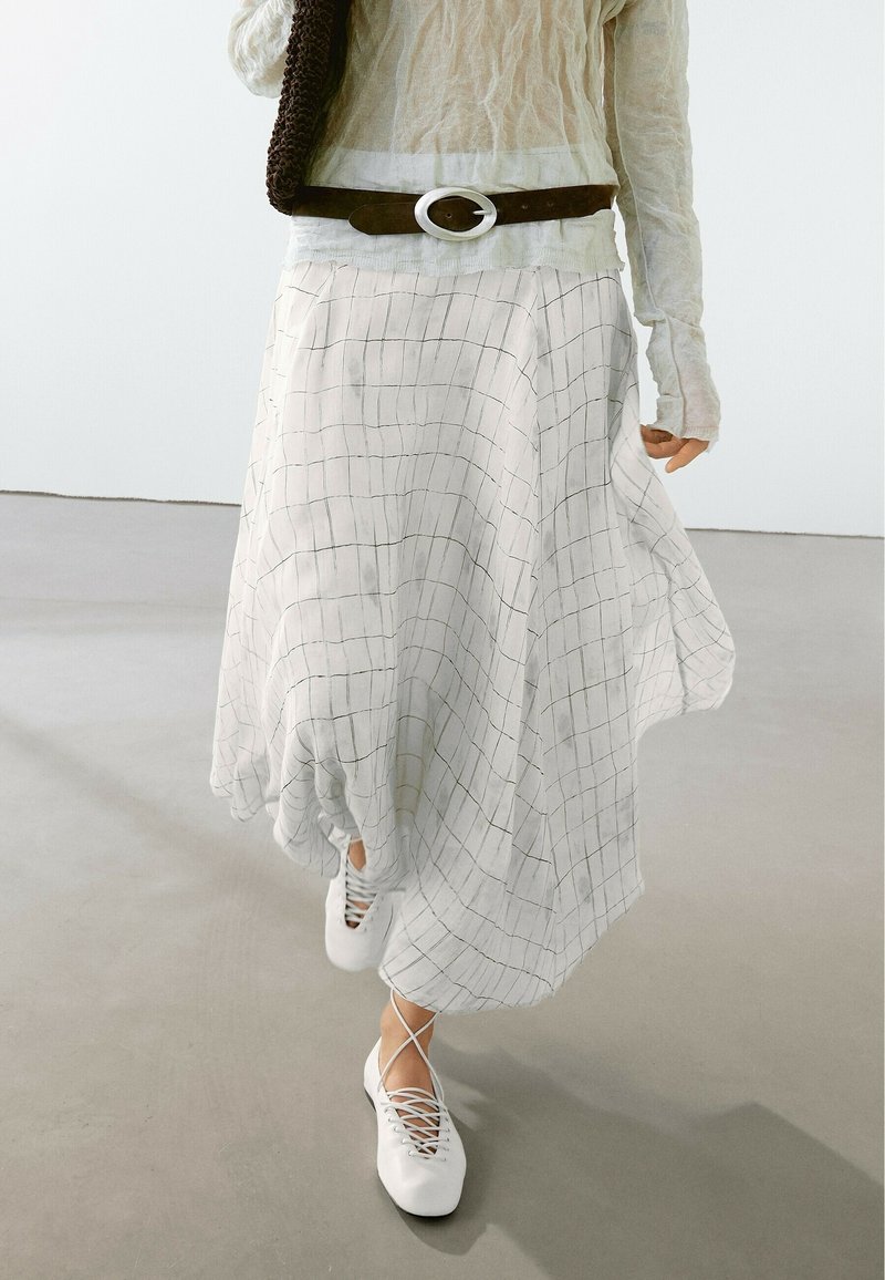 Personne portant une jupe fluide à motif quadrillé blanc, un haut beige à manches longues, une ceinture marron, un sac marron et des ballerines blanches à lacets marchant à l'intérieur.