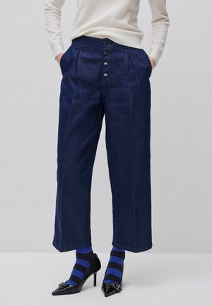 Femme portant un pantalon large court bleu foncé avec des boutons à l'avant, des chaussettes rayées bleu et noir, et des escarpins noirs à bout pointu.