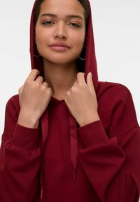 Sudadera burdeos con cordón, textura suave, corte oversize y puños acanalados. La capucha cubre la cabeza, ofreciendo una cobertura y calidez completas.