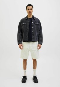 Veste en denim sombre avec poches avant, fermeture à boutons, portée sur une chemise noire. Un short clair, des chaussettes blanches et des chaussures noires complètent la tenue.